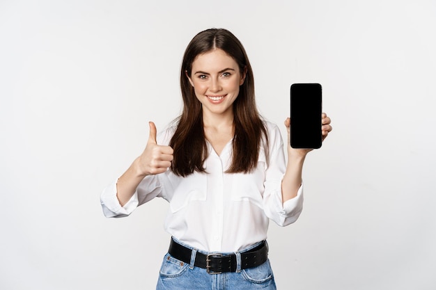 엄지손가락을 치켜들고 웃고 있는 여성, 추천 앱, 휴대폰 화면, 스마트폰의 빈 인터페이스, 흰색 배경에 서 있습니다.