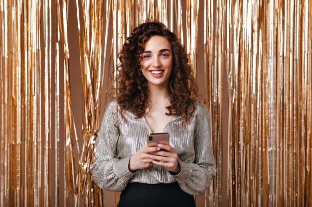 Smiling woman in silver blouse holding smartphone on golden background