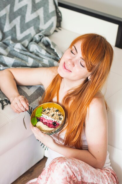 Smiling woman sitting on floor with closed eyes holding bowl of oatmeals