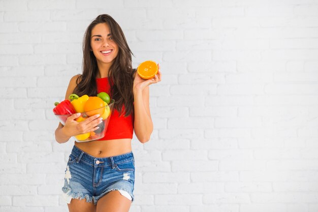 Smiling woman standing against wall holding fresh vegetable and fruits