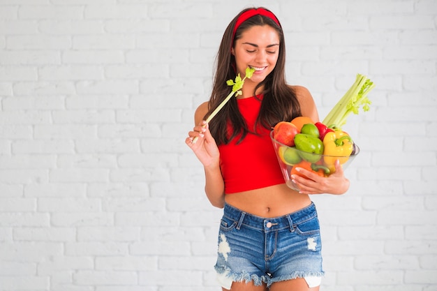 Smiling woman standing against wall holding fresh vegetable and fruits