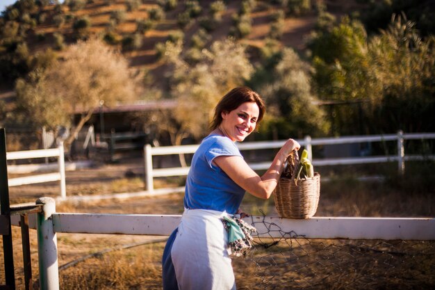 수확 한 야채 바구니를 들고 목장 근처에 서 웃는 여자