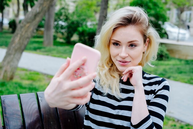 Donna sorridente che prende selfie con lo smartphone mentre sedendosi all'aperto su un banco nella cima a strisce