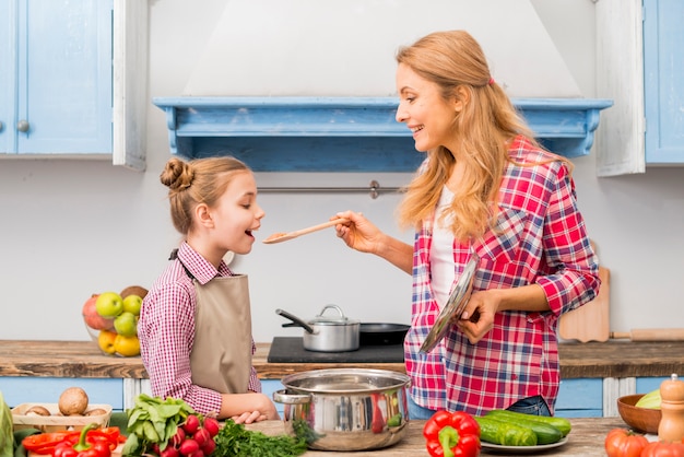 Smiling woman tasting a food to her daughter with a wooden spoon in the kitchen