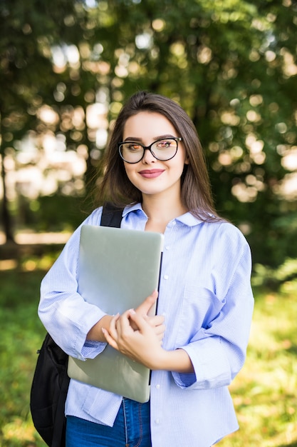 Улыбающаяся женщина в прозрачных очках стоит со своим ноутбуком в парке