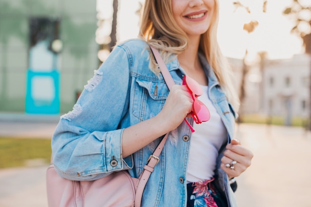 smiling woman walking in city street in stylish denim oversize jacket, holding pink leather backpack