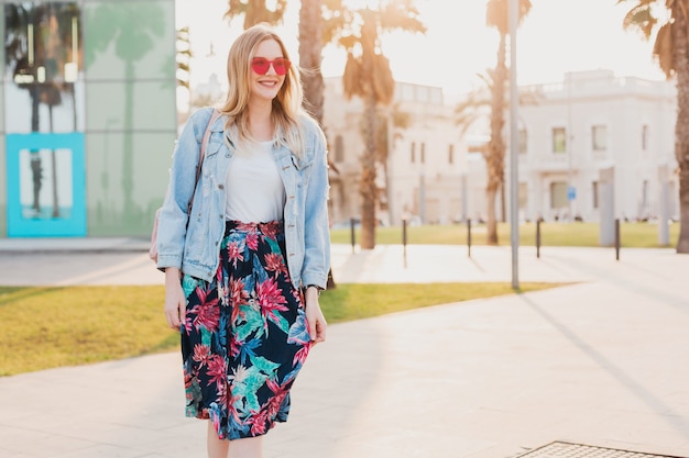 smiling woman walking in city street in stylish printed skirt and denim oversize jacket wearing pink sunglasses