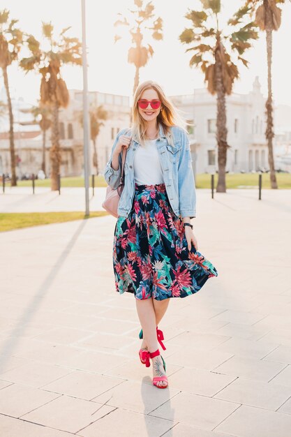 smiling woman walking in city street in stylish printed skirt and denim oversize jacket wearing pink sunglasses