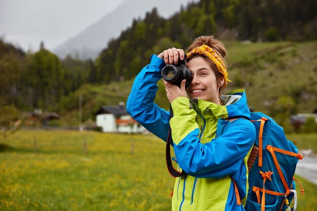 Smiling woman wanders in beautiful mountain countryside, keeps camera near eye, makes photo dressed in casual jacket