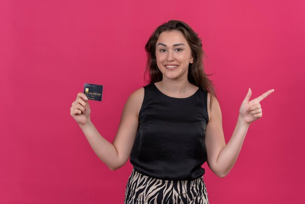 Smiling woman wearing black undershirt holding a bank card and point to side - on pink wall