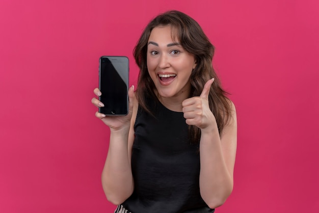 Улыбающаяся женщина в черной майке держит телефон и показывает ей палец на розовой стене