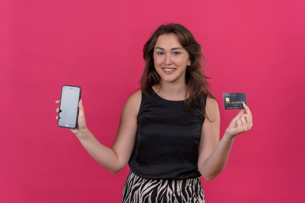 Smiling woman wearing black undershirt holding a phone and bank card on pink wall