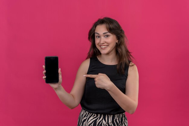 Smiling woman wearing black undershirt holding a phone and point the phone on pink wall