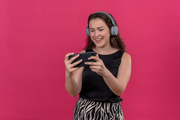 Smiling woman wearing black undershirt listen music from headphones on pink wall