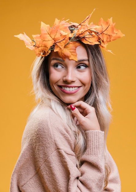 Smiling woman wearing dry maple leaves tiara standing against yellow wall looking away