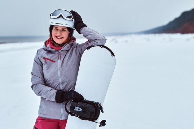 Smiling woman wearing warming sportswear posing with a snowboard on a snowy beach