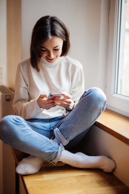 窓辺に座っていると、自宅のスマートフォンでメッセージを書く白いセーターで笑顔の女性