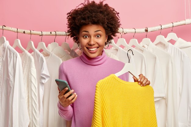 Smiling woman with Afro hair, uses mobile phone app for paying online, buys new yellow sweater, stands behind clothing rail