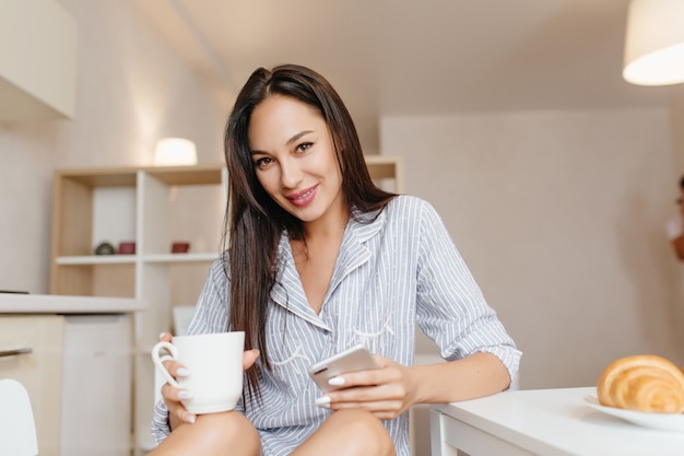 朝食時にスマートフォンでキッチンに座っている黒髪の笑顔の女性