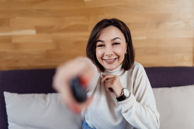 ブルネットの髪を持つ笑顔の女性は彼女のモダンな明るいフラットでテレビをオンにします