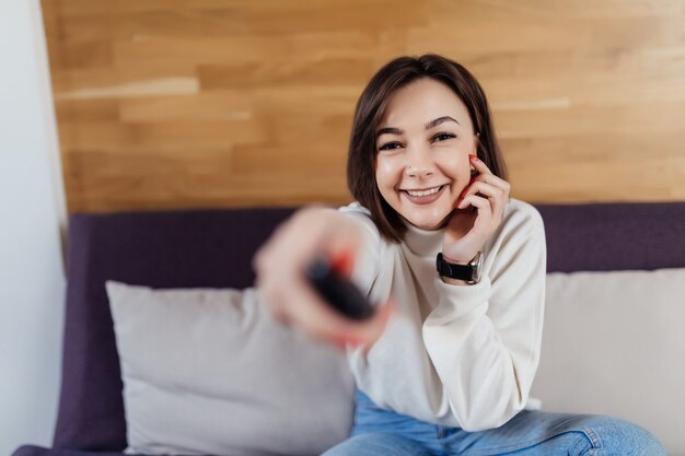 Smiling woman with brunette hair is turning on tv-set in her modern bright flat