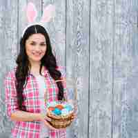 Free photo smiling woman with bunny ears on head showing easter eggs basket against wooden backdrop