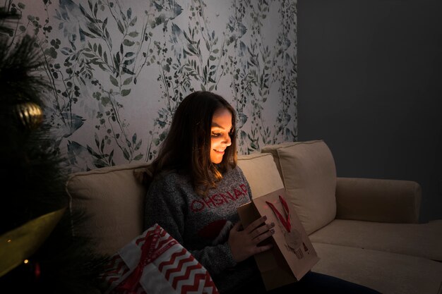 Smiling woman with gift packet on settee near Christmas tree