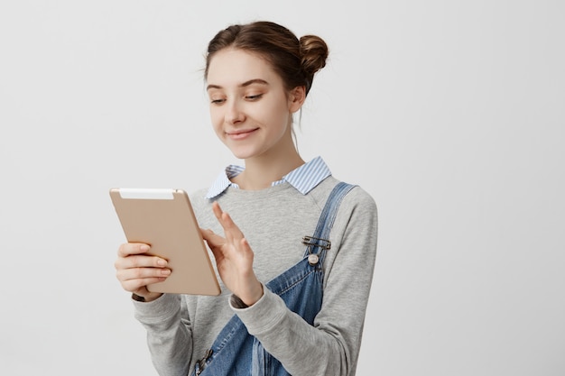 Smiling woman with hair tied in double buns looking at screen of modern device. Pleased female cutie typing message to her boyfriend using tablet. Relationships concept