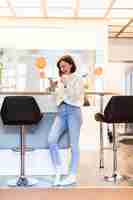 Free photo smiling woman with phone standing in panoramic kitchen with bright walls high table and bar chairs