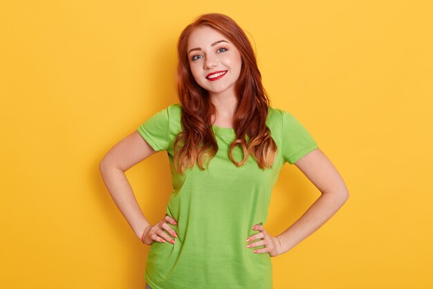 Smiling woman with red hair and bright lips posing isolated, holding hands on hips, looking smiling directly at camera