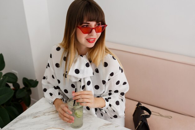 Smiling woman with red lips holding beverage, sitting on sofa on cafe