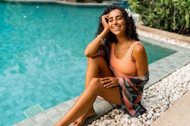 Smiling woman  with wavy hairs chilling near swiming pool  Tropical stylish outfit Plumeria flower in hairs