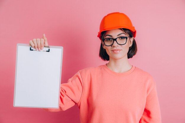 Smiling woman worker builder hold white sign board blank against pink background. Building helmet.