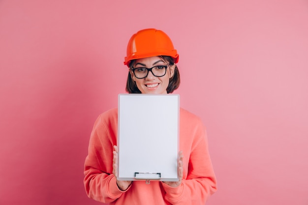 Smiling woman worker builder hold white sign board blank against pink background. Building helmet.