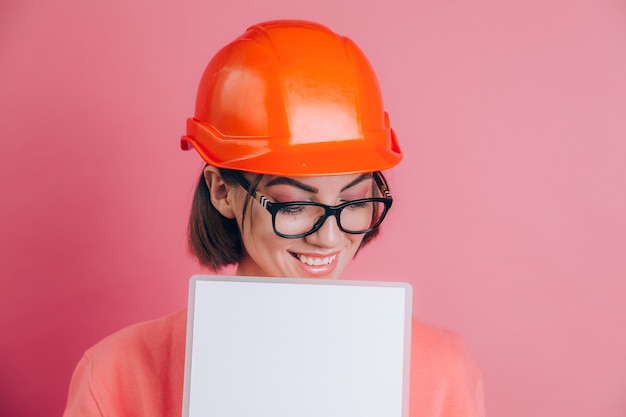 笑顔の女性労働者ビルダーは、ピンクの背景に白い看板を空白に保持します。建物のヘルメット。
