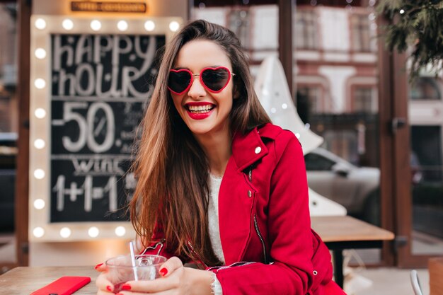 Улыбающаяся замечательная девушка в стильных солнцезащитных очках проводит утро выходного дня в летнем кафе