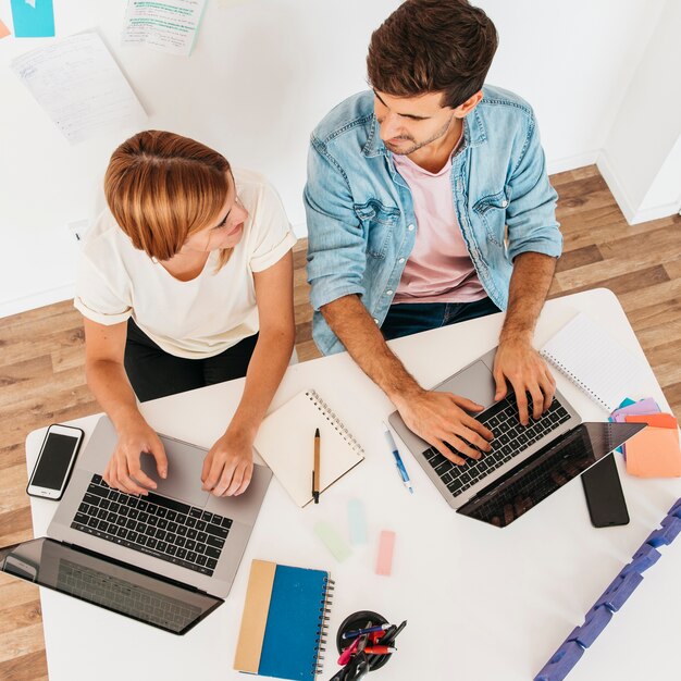 Smiling working male and female sitting at workplace and using laptops looking at each other