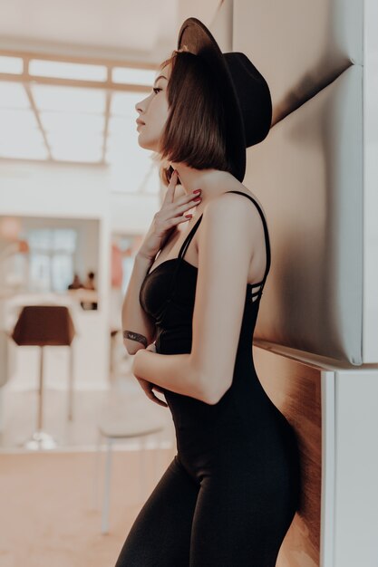 黒のドレスと帽子の高級マンションの壁の近くに立って笑顔の若い大人のブルネットの女性