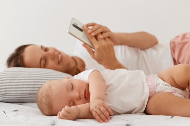 Smiling young adult female with dark hair using cell phone for checking social networks or browsing internet while lying on bed with infant sleeping baby.