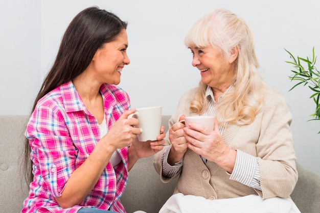 Smiling young adult and senior mother holding cup of coffee looking at each other