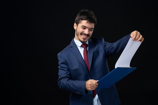 Smiling young adult in suit holding documents and checking statistics in it on isolated dark wall