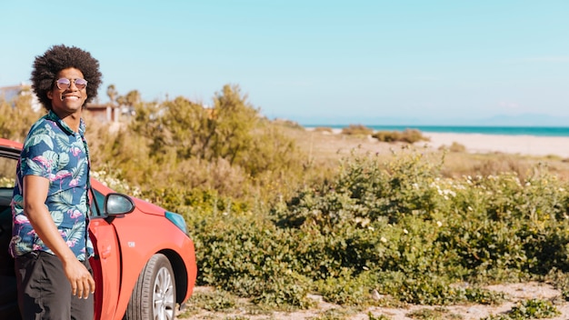 Giovane uomo afroamericano sorridente che sta vicino all'automobile sulla spiaggia