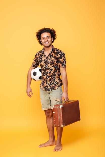 Улыбающийся молодой африканский мужчина держит чемодан и футбол