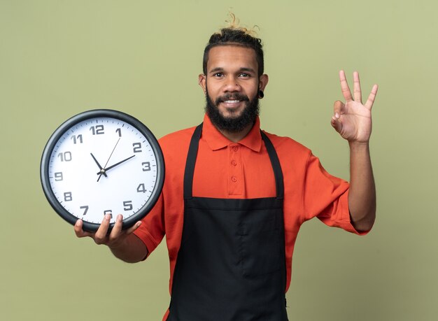 smiling young afro-american barber wearing uniform holding clock looking at camera doing ok sign isolated on olive green background