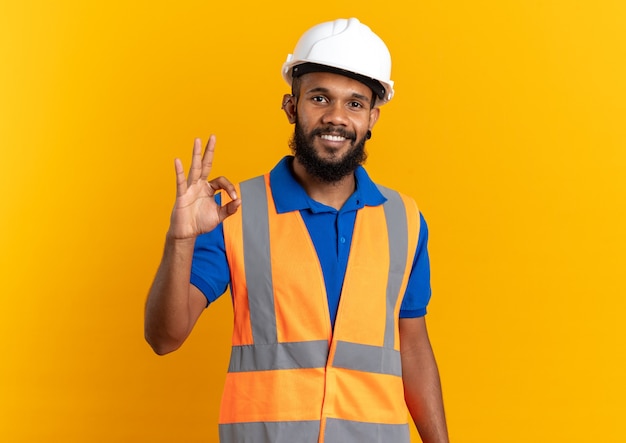 Sorridente giovane costruttore afroamericano uomo in uniforme con casco di sicurezza che gesturing segno ok isolato sulla parete arancione con spazio copia