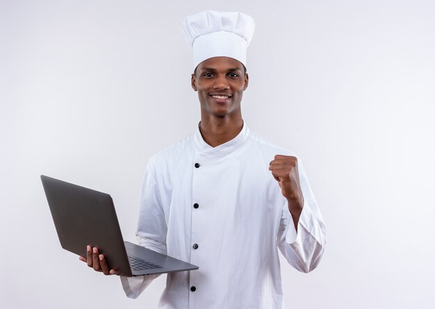 Smiling young afro-american cook in chef uniform holds laptop and keeps fist up on isolated white background with copy space