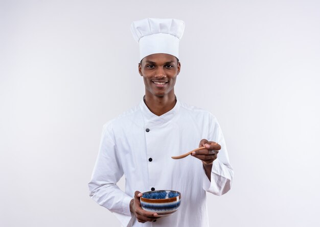 Smiling young afro-american cook in chef uniform holds and points at bowl on isolated white background with copy space