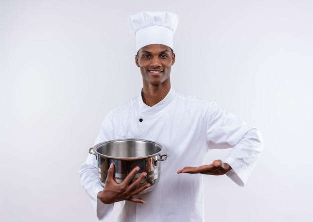 シェフの制服を着た笑顔の若いアフリカ系アメリカ人料理人は鍋を保持し、コピースペースで孤立した白い背景に手をまっすぐに保ちます