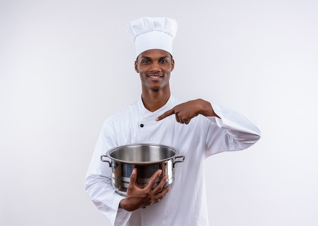 Smiling young afro-american cook in chef uniform holds saucepanand points at saucepan on isolated white background with copy space