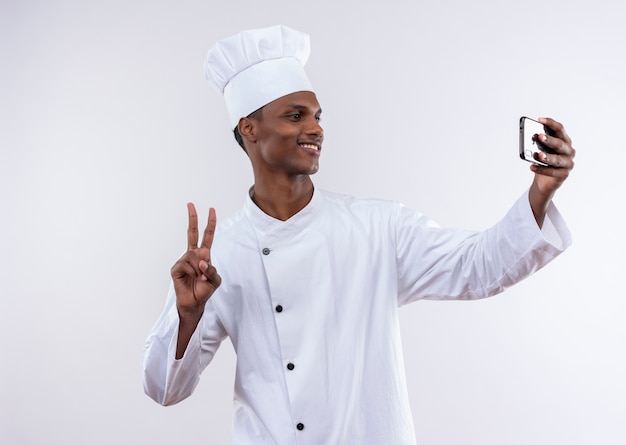 Smiling young afro-american cook in chef uniform looks at phone and gestures victory sign on isolated white background with copy space